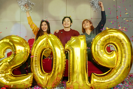 Group of cheerful asian young people friends sitting on red sofa carrying balloon gold colored numbers and throwing confetti on the party. New 2019 Year is coming Celebrate. Bokeh light soft effect.の写真素材