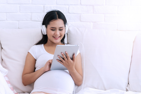 Attractive happy pregnant woman relaxing while listening to music on headphones from tablet in her hand sitting on white sofa at home.の写真素材