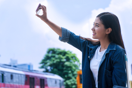 Traveler young woman using smartphone. Shooting themselves "Selfie" in train station. Attractive girl taking selfie by mobile phone on outdoor at railway station.の写真素材