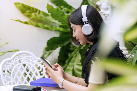 Concept relax time. Attractive back of young women wearing white headphones enjoy listening music from the mobile while sitting in her garden. Front have blurred leaves.の写真素材