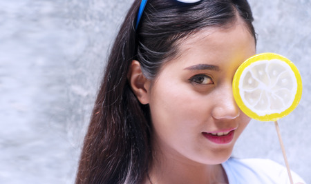 Cheerful young woman covered her eye with bright fruit shaped lollipop over gray background with copy space.の写真素材