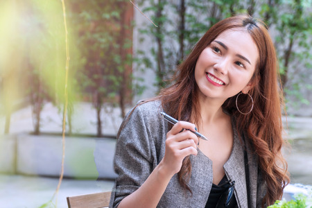 Charming young woman smiling look like thinking about something to use her idea with copy space. Attractive business woman sitting in coffee shop smile and thinking looking to the side with pen.の写真素材