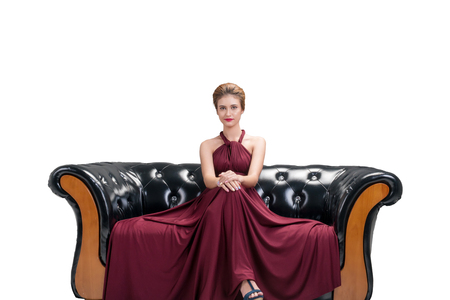 Attractive young woman wear red dress fashion posing and sitting on black sofa looking at camera on isolated white background. Beauty and luxurious conceptの写真素材