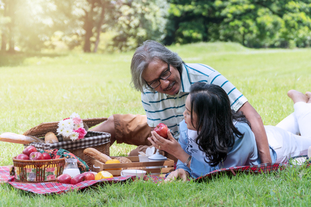 Elderly lifestyle concept. Happiness senior couple relax together in the public park. Husband embracing and holding red apple for his wife with picnic on green grass.の写真素材