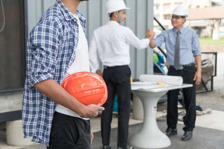 Close up of builder holding orange hardhat or helmet at construction site while two construction engineers finished working shaking hands. Concept of success and teamwork.の写真素材