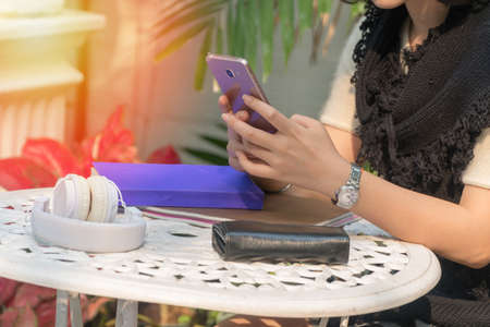 Hand of young woman hold her smartphone with white headphones and books sitting and relaxing outdoor in the garden.の写真素材