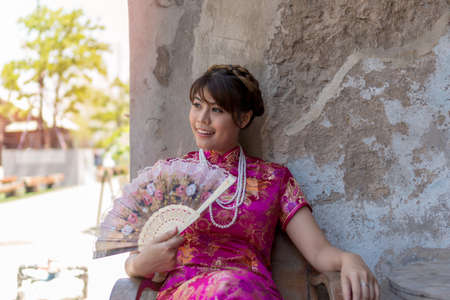 Portrait beautiful young woman wear cheongsam deep pink dress holding a fan looking something....
