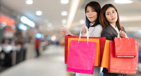 Happiness, consumer, sale and people concept. Attractive two young woman holding shopping bags looking at camera showing thumbs up on shopping mall background with copy space.の写真素材