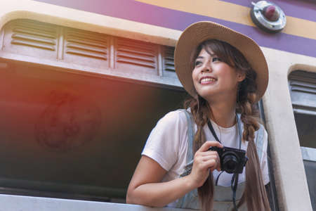 Attractive cheerful asian young woman wearing hat from window train holding camera in hand. Young girl leans out of the train window for looking interest place or tourist location.の写真素材