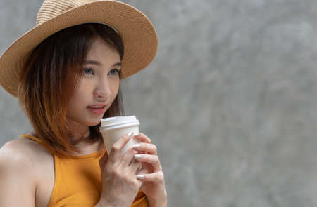 Attractive women standing hold cup of coffee on concrete walls background with copy space. Gorgeous woman wearing casual clothes and hat loves drinking coffee that make her fresh.の写真素材