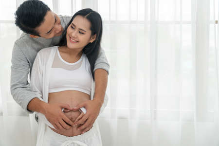 Husband embracing his wife making heart shape on the pregnant belly with their hands. Happy couple...
