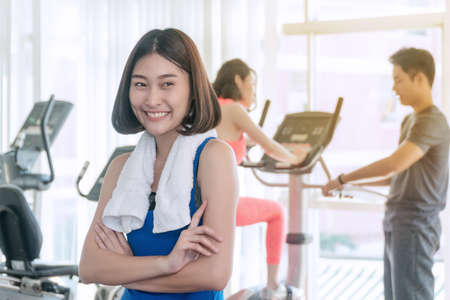 Cheerful sports woman standing arms crossed in fitness gym. Attractive woman wearing blue sportswear smiling happily at gym in front of elliptical machines.の写真素材