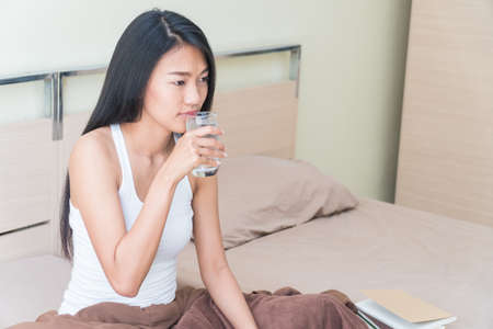 Beautiful young woman sitting in bed holding a glass of water to drink.の写真素材