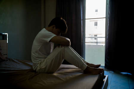 Melancholy young man barefoot sitting at the bed embracing his knees, his head down, Feeling problem...