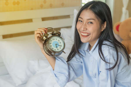 Beautiful young woman sitting on the bed holding alarm clock in hand.の写真素材