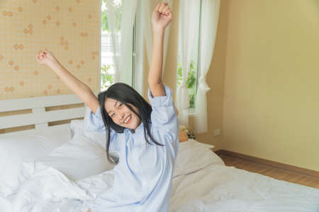 Asian beautiful girl wakes up, stretches her arms in a white bed in the bedroom.の写真素材