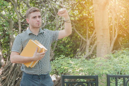 Smart young man standing holding a yellow book in the garden under the tree with the sun shining.