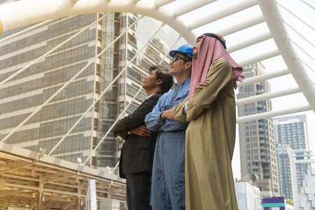 Three men standing arms folded together looking up. Middle-aged businessman,Young engineers and Arab businessmen standing in a row looking in same direction In modern urban background.Teamwork concept.の写真素材