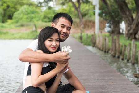 The couple sitting together on the bridge beside the pond water. young man sitting hugging girlfriend holding flowers in hand on natural background. Concept relaxationの写真素材