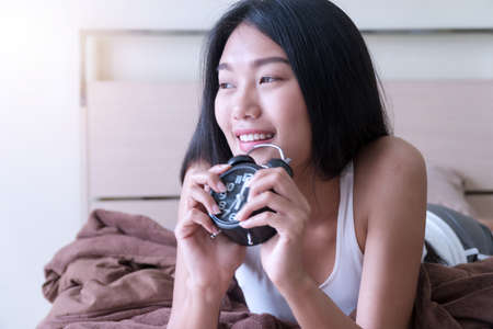 Smiling young woman waking up holding alarm clocks on the bed in the morning. Lifestyle concept.の写真素材