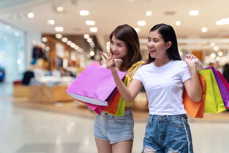 Cheerful excited two young woman holding colorful shopping bags pointing fingers to something on shopping malls background. Happiness, consumerism, sale and people concept.の写真素材