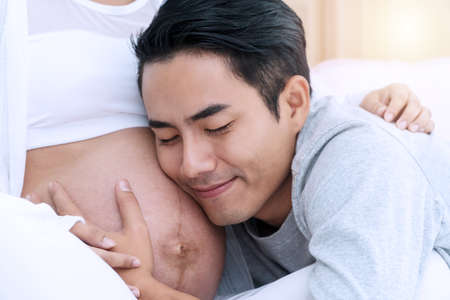 Husband listening the belly of his wife near the window in the home. Handsome young man listen the belly of his lovely pregnant wife standing in the room. Happy family pregnancy, expectation.の写真素材