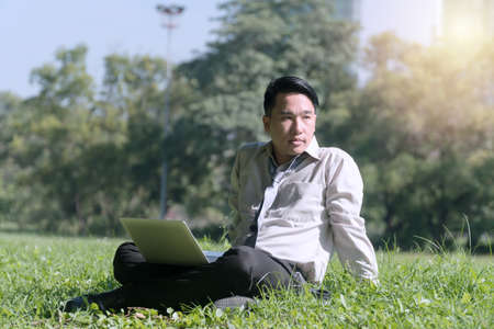 Businessman with laptop sitting on green grass working and looking at something in the park. Lifestyle relaxation concept.の写真素材