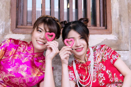 Cheerful young woman wear cheongsam dress looking at camera on Chinese atmosphere background. Two young girl holding red heart shape in their hands with close eye.の写真素材