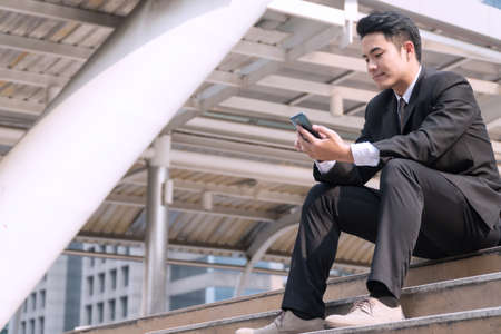Handsome young businessman wearing black suit sitting on stair using smartphone in modern city background. Professional young man holding cellphone in his hand working online on outdoor.の写真素材