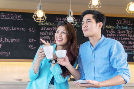 Happy young couple holding cup of coffee in cafÃ©. Cheerful young woman pointing fingers to her boyfriend see something in outdoor coffee shop.の写真素材