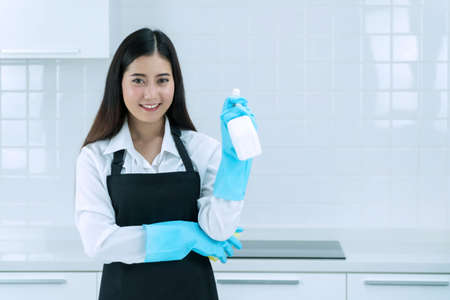Housekeeper concept. Professional young woman wearing aprons and rubber protective blue gloves holding bottle cleaners in the kitchen. Housewife smile hold bottle spray work clean at home.の写真素材