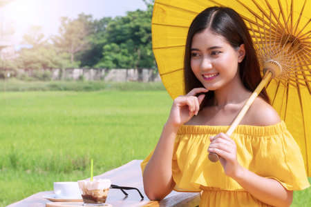 Joyful smile young women wear yellow dresses holding yellow umbrella sitting relax with camera, cup of coffee and cakes looking at something in the rice field. Concept lifestyle relaxの写真素材