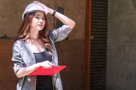 Engineer young woman wearing white helmet stand at outdoor holding the folder with some documents on site job. Architect women wearing construction hard hat or helmet checking work in progress at construction site.の写真素材