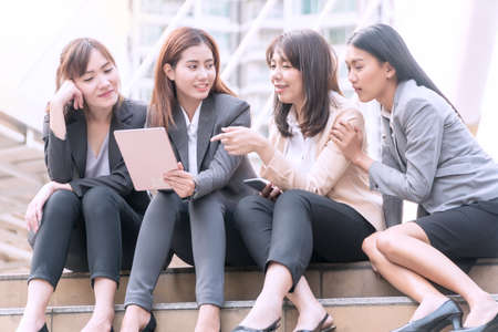 Group of businesswoman sitting in the shade on the stairs,looking at digital tablet in a modern city. Beautiful young woman wearing the business suit using tablet and laughing happily.の写真素材