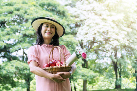Elderly lifestyle concept. Cheerful smiling senior women 50s with hat while holding fruit basket and bouquet of flower in her hands looking at camera in the public park.の写真素材