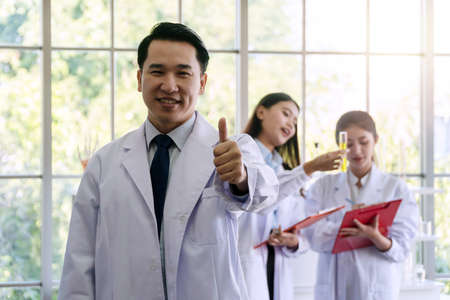 Researchers man in white coat showing thumbs up over busy researchers team in the laboratory looking at camera.の写真素材