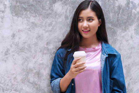 Attractive happy smile young woman standing hold cup of coffee on concrete walls background with...