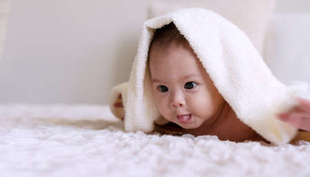 Cheerful cute baby looking at camera under white blanket looking at something. Innocence baby crawling on white bed with towel on his head at home.の写真素材