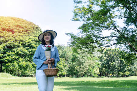 Elderly lifestyle concept. Cheerful smiling senior women 50s with hat while holding fruit basket and bouquet of flower in her hands looking at camera in the public park.の写真素材