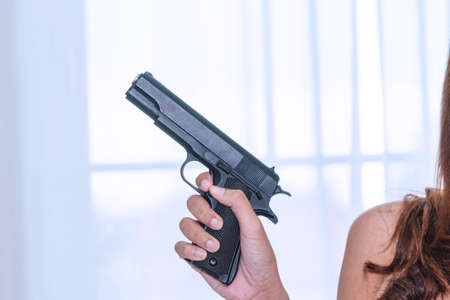 Woman holding gun in her hand at home. Focus hand of young women using black gun protection from danger gesture killer.の写真素材