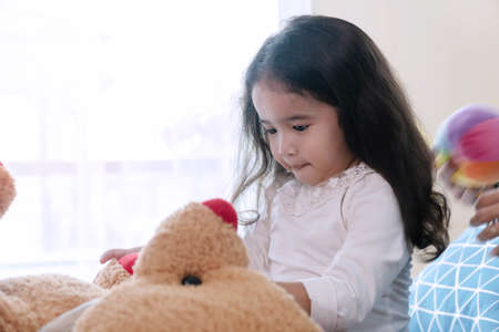 Cheerful cute little girl playing big brown teddy bear sitting on bed in bedroom. Lifestyle child relaxation conceptの写真素材