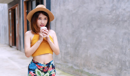 Attractive women standing hold cup of coffee on concrete walls background with copy space while looking at something. Gorgeous woman wearing casual clothes and hat loves drinking coffee make her fresh.の写真素材
