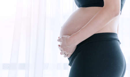 Close up of abdomen pregnant young woman standing beside windows in the bedroom. Body pregnancy mother use hands holding her belly at home. Lifestyle prenatal concept.の写真素材