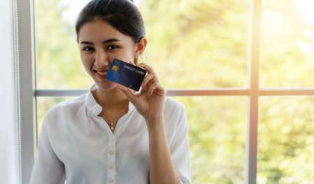 Attractive smiling happy young woman holding blank card in her hands at front windows in room with copy space. Cheerful girl showing mock up bank card while looking camera. Payment blanking concept.の写真素材