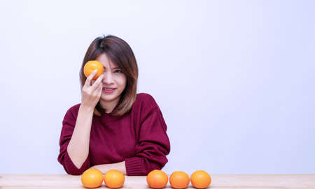Healthy food concept. Cheerful young woman holding orange covered her eyes while sitting on table looking at camera on white background.の写真素材