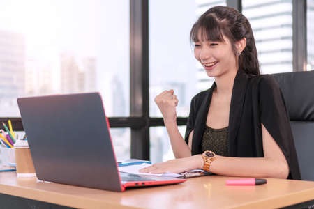 Accountant young woman sitting in coworker while looking at laptop, She raised fist up feeling cheer success her project. Joyful businesswoman with notebook on desk in workplace successful her job.の写真素材