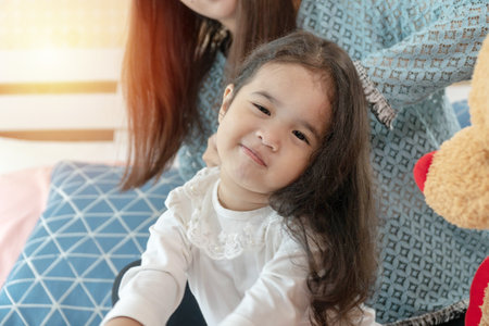 Little cute girl sitting on the bed while her mother combing daughter's hair together. Cheerful adorable childhood feel comfort with mom making hairstyle at home.の写真素材