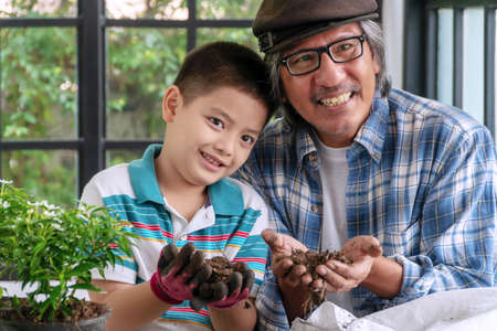 Activity little cute boy and his grandfather holding dry ground in their hands on greenhouse.Happy smile grandfather teaching his grandson growing tree while hold soil in their hands together at home.の写真素材