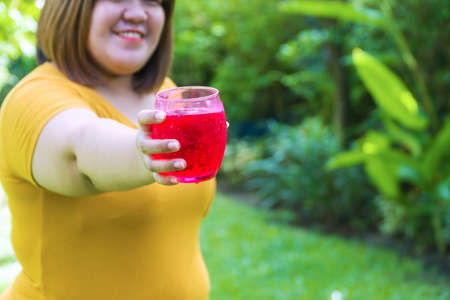 Fat young woman holding a glass of red nectar with ice soda in her hand while standing in the garden. Eat a lot. Not good for health. Overweight obesity Unhealthy concept.の写真素材
