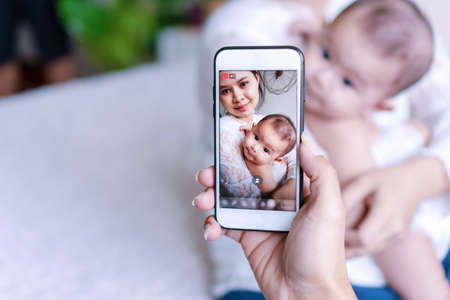 Hand of husband man holding smart phone while taking his son and wife together in the room. Picture cheerful young woman with his baby on touch screen phone while live on social. Technology concept.の写真素材